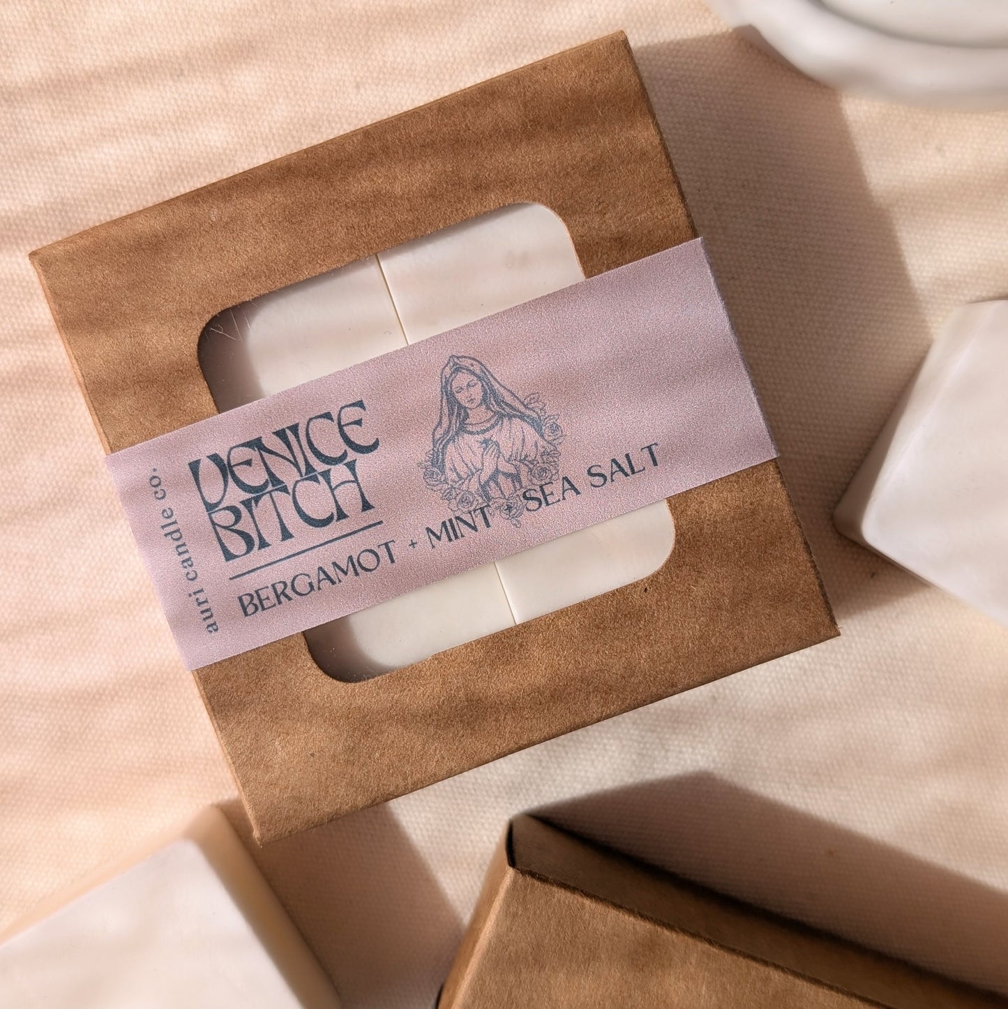 A packaged product labeled 'Venice Bitch Bergamot + Mint + Sea Salt' with a graphic of a woman's face, suggesting a connection to the fragrance or brand theme.